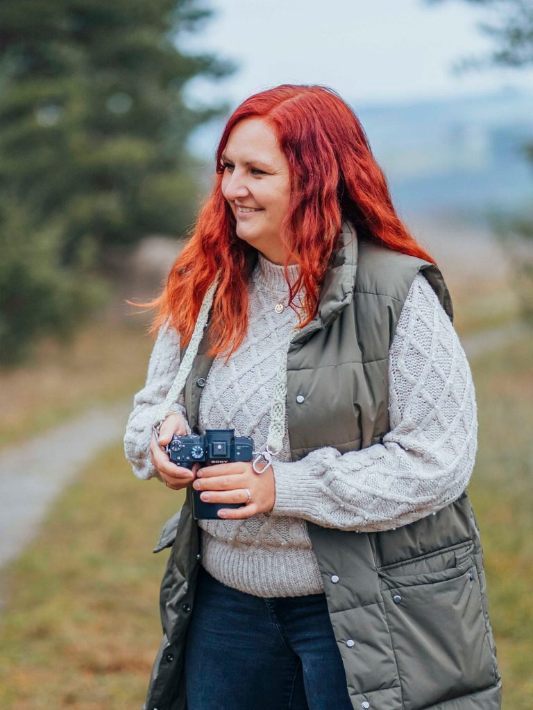 Familienfotografin in Main Spessart & Umgebung Frau mit rotem Haar, die in einem Waldgebiet mit Kamera unterwegs ist.