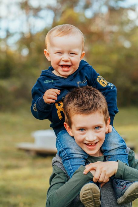 Ein fröhlicher Junge trägt ein lachendes Kleinkind auf den Schultern im Freien.