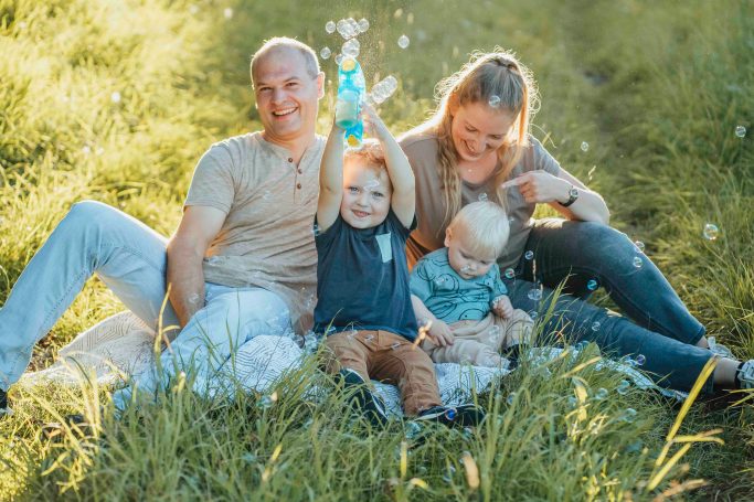 Familie sitzt im Gras, zwei Kinder spielen, eine Person hält ein Wasserflasche.